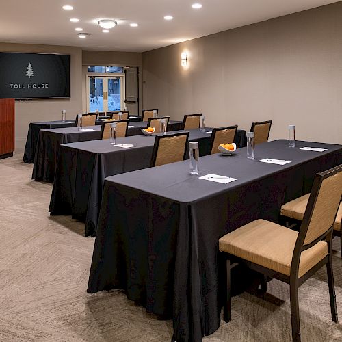 The image shows a small conference room setup, with tables and chairs arranged facing a podium and a screen displaying 