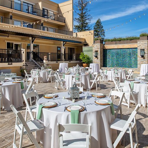 An outdoor event setup with round tables covered in white tablecloths and green napkins, white chairs, a fountain wall, and a multi-story building.