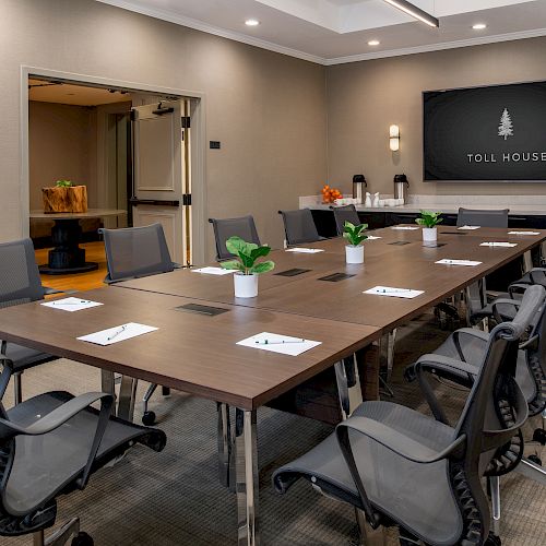 This image shows a modern conference room with a long table, ergonomic chairs, potted plants, and a screen displaying 