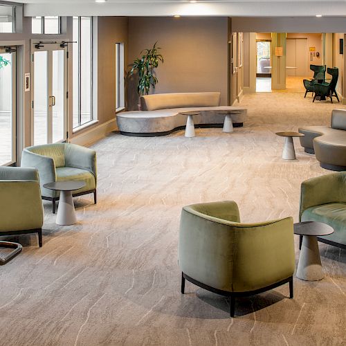 A modern, spacious lobby with green and gray armchairs, small tables, large windows, and indoor plants on beige carpeting.