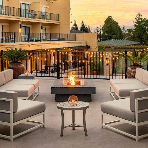 An outdoor seating area with a fire pit, surrounded by chairs and tables, lit by string lights, with a building and trees in the background.