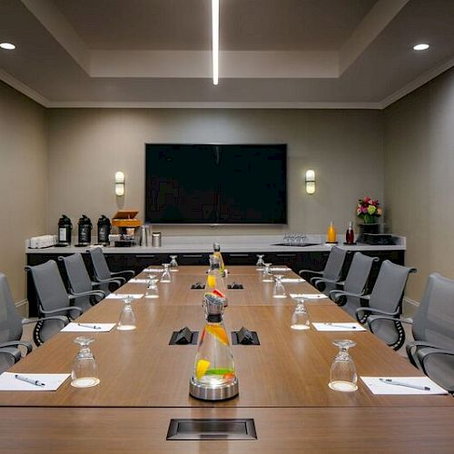 A modern conference room with a long table, chairs, notepads, and water glasses, with a large screen on the wall, and beverages on the side table.
