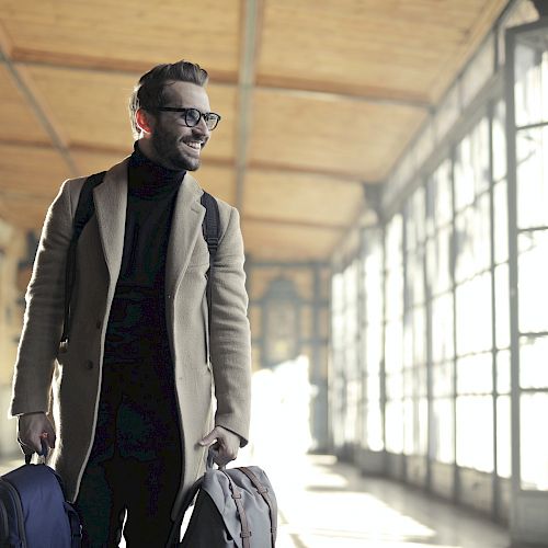 A man with glasses, wearing a coat, is walking in a bright hallway with two bags, smiling and looking to the side.