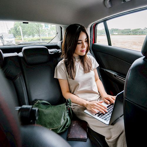 A person is sitting in the backseat of a car, working on a laptop with a green bag and a phone beside them, and looking focused.