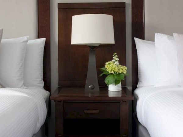 Two neatly made beds with white linens and pillows, separated by a nightstand holding a lamp and a small vase with flowers.