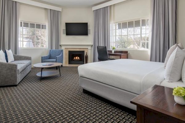 A modern hotel room with a bed, sofa, coffee table, armchair, fireplace, desk, and TV, complemented by large windows with curtains.