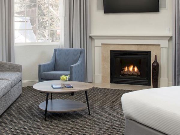The image shows a cozy living space with a sofa, armchair, coffee table, fireplace, TV, large window, and bed, all in a neutral color palette.
