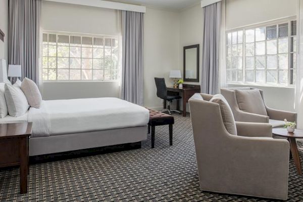 This image shows a hotel room with a large bed, two armchairs, a small round table, a desk, and large windows with curtains.