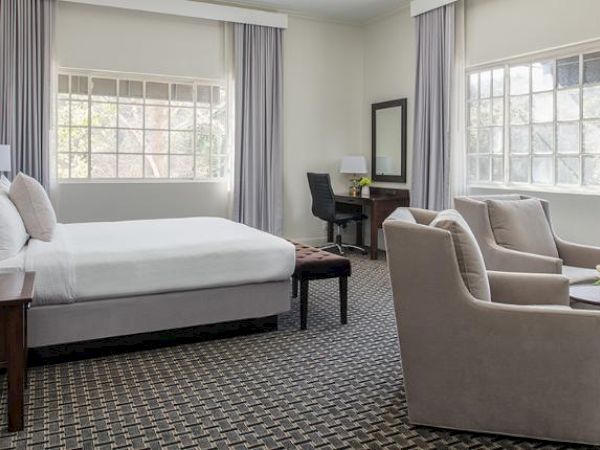 This image shows a hotel room with a large bed, two armchairs, a small round table, a desk, and large windows with curtains.