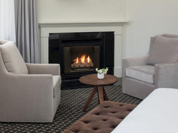 A cozy room with two upholstered chairs, a small round coffee table, and a fireplace. A bed and an ottoman are also visible in the scene.