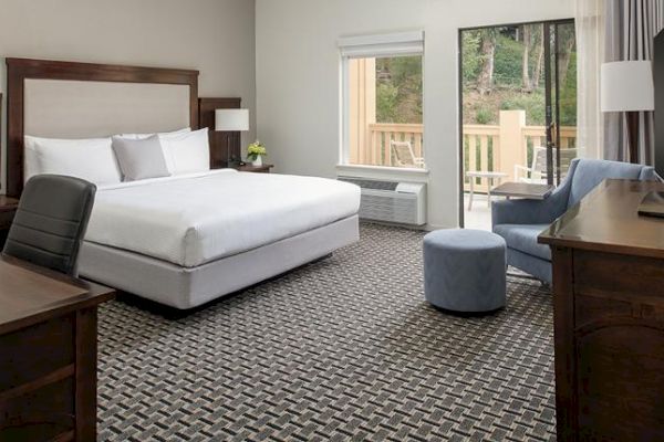 This image shows a hotel room with a large bed, a desk with a chair, an armchair, a TV on a dresser, and a window with a view of greenery outside.