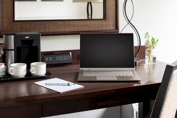 A desk with a laptop, coffee maker, cups, paper, and a pen, set against a mirror and a lamp, with flowers in a vase on the side.