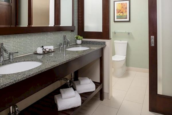 A modern bathroom with double sinks, mirrors, a toilet, tiled floors, towel storage, and a framed picture on the wall.