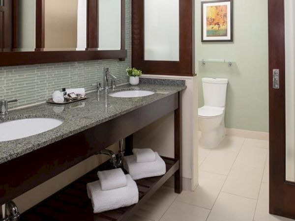A modern bathroom with double sinks, mirrors, a toilet, tiled floors, towel storage, and a framed picture on the wall.