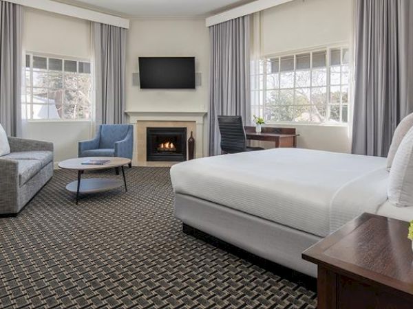 A spacious hotel room features a bed, a gray sofa, a blue chair, a TV over a fireplace, large windows with curtains, and a desk with a chair.