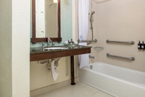 A bathroom with a sink, mirror, and bathtub with grab bars and mounted dispensers.