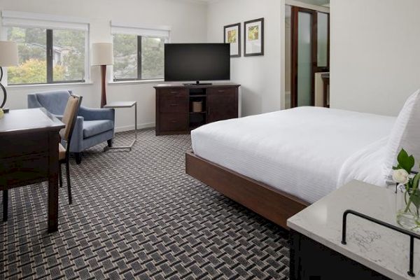 A neatly furnished hotel room features a bed, desk, chair, TV, and large windows allowing natural light, with decor including framed pictures.