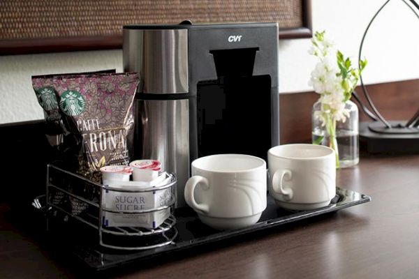 The image shows a coffee station with a coffee maker, two coffee mugs, sugar packets, and a bag of Starbucks coffee. A small floral arrangement is beside it.