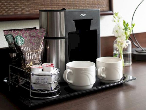 The image shows a coffee station with a coffee maker, two coffee mugs, sugar packets, and a bag of Starbucks coffee. A small floral arrangement is beside it.