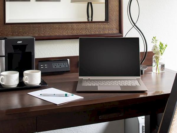 A desk setup with a laptop, coffee machine, two cups, a notepad, a pen, a power outlet, and a small flower vase against a white wall with a mirror.