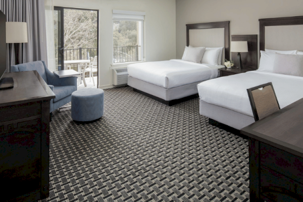 The image shows a hotel room with two beds, a desk, a TV, a blue chair, an ottoman, and large windows with a view of trees outside.