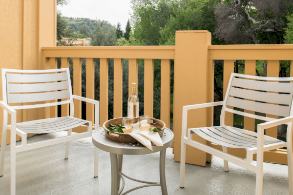 Two white chairs on a patio with a small table in between. The table holds a tray with snacks and a bottle, surrounded by wooden railings and greenery.