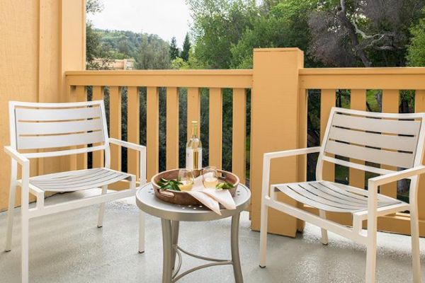 A balcony scene with two white chairs, a small table with a bottle, glasses, a tray, and napkins. Greenery and trees are visible in the background.