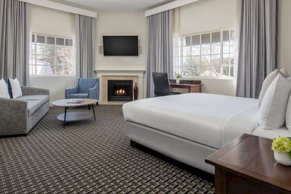 A modern hotel room features a bed, a sofa, a fireplace, a flat-screen TV, a desk, and large windows with curtains, exuding a cozy yet elegant ambiance.