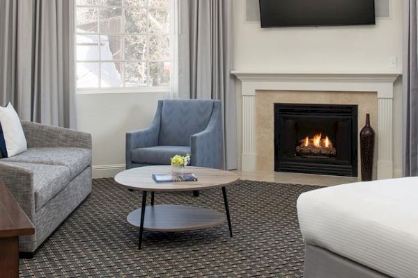 A cozy living room with a grey sofa, blue armchair, round coffee table, fireplace, wall-mounted TV, and large window with grey curtains.