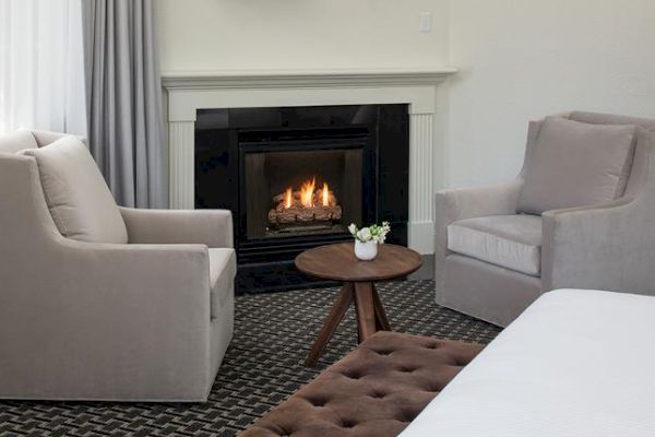 A cozy room with two beige armchairs, a small round table with a flower vase, a lit fireplace, and a bed with a tufted ottoman.