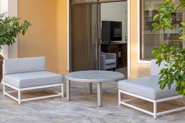 A cozy outdoor seating area with two gray cushioned chairs, a round table, potted plants, and a view of the indoors through a glass door.