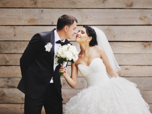 A bride and groom in wedding attire share a moment, with the bride holding a bouquet, against a wooden background.