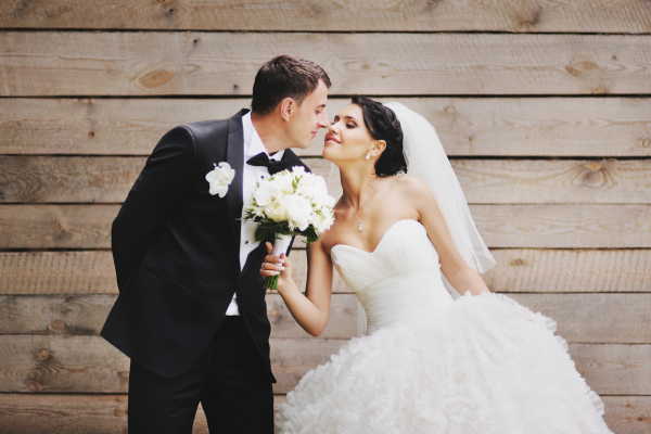 A bride and groom in wedding attire share a moment, with the bride holding a bouquet, against a wooden background.