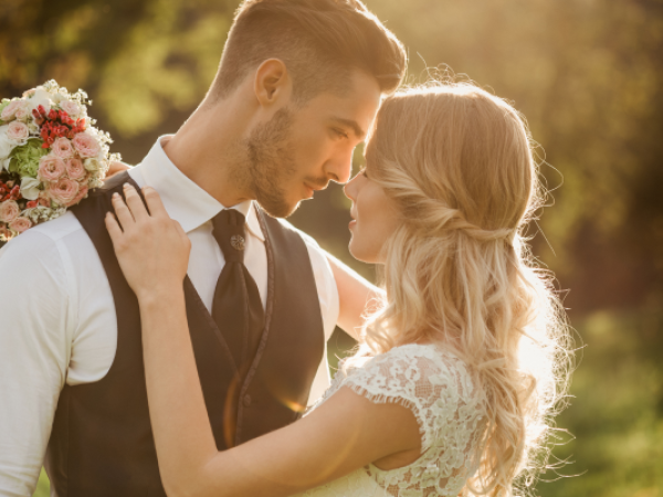 A couple is embracing outdoors, forehead to forehead, with the woman holding a bouquet of flowers in her right hand.