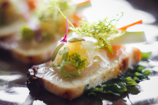 The image shows a gourmet dish featuring slices of fish topped with various garnishes and served on greens, presented artistically on a plate.