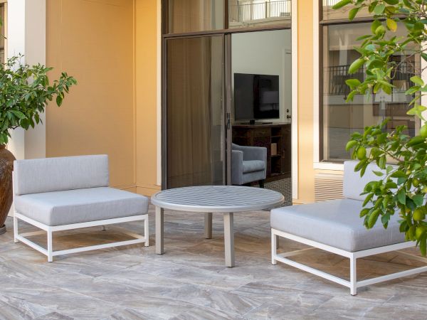 The image shows an outdoor seating area with two gray cushioned chairs, a round table in between, and some greenery in the background and foreground.