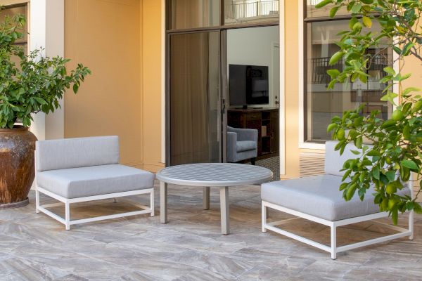 The image shows an outdoor seating area with two gray cushioned chairs, a round table in between, and some greenery in the background and foreground.