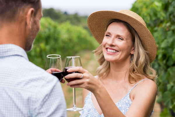 A woman in a straw hat smiles, clinking wine glasses with a person in a vineyard.