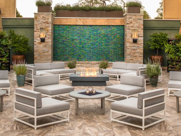 An outdoor seating area with modern lounge chairs, a central fire pit, a water feature on a tiled wall, and surrounding plants.