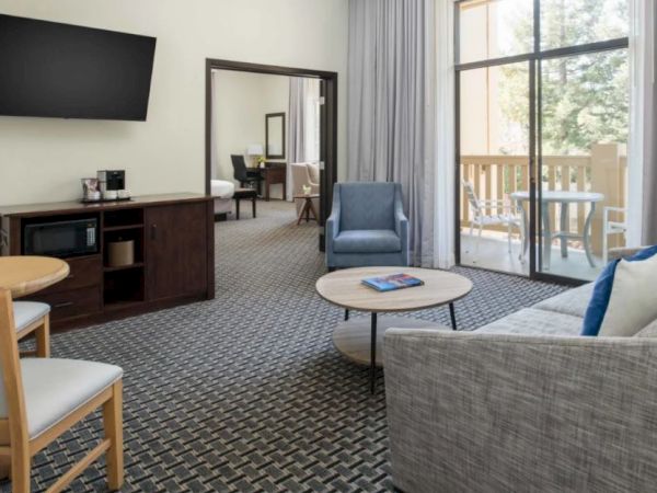 The image shows a modern living room with a sofa, armchair, round coffee table, flat-screen TV, small dining table, and a balcony with outdoor seating.
