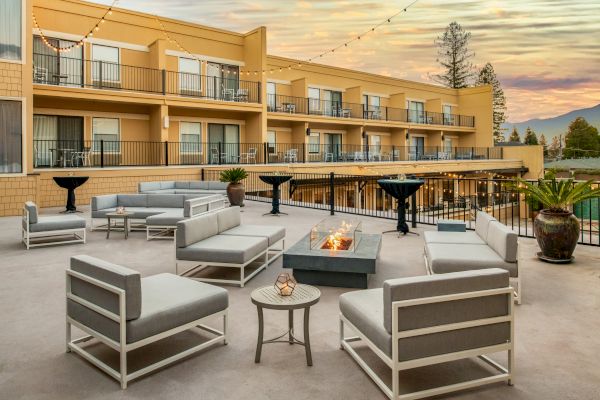 An outdoor lounge area with modern furniture, a fire pit, and potted plants. The setting is against a building with balconies and sunset views.