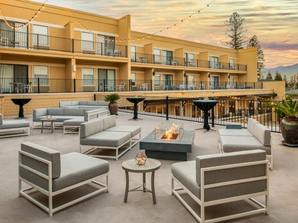 An outdoor lounge area with modern furniture, a fire pit, and potted plants. The setting is against a building with balconies and sunset views.