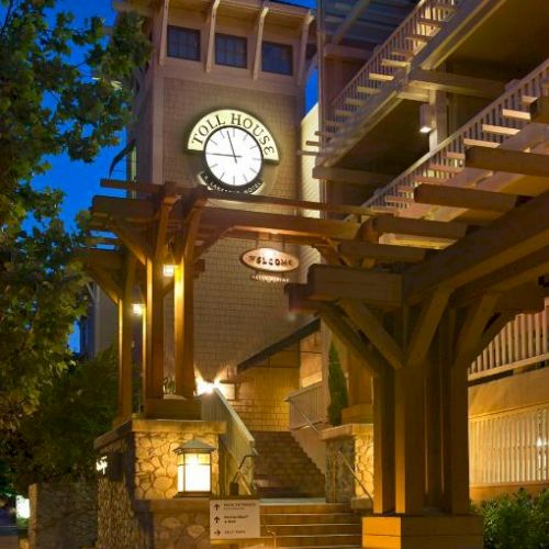 The image shows a building with illuminated wooden beams, stone accents, and a clock tower at the front, under a deep blue evening sky.