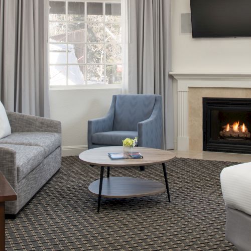 A cozy room with a fireplace, gray sofa, blue armchair, round coffee table, bed, and TV mounted above the fireplace, near a window.