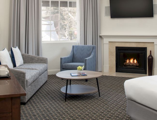 A cozy room with a fireplace, gray sofa, blue armchair, round coffee table, bed, and TV mounted above the fireplace, near a window.