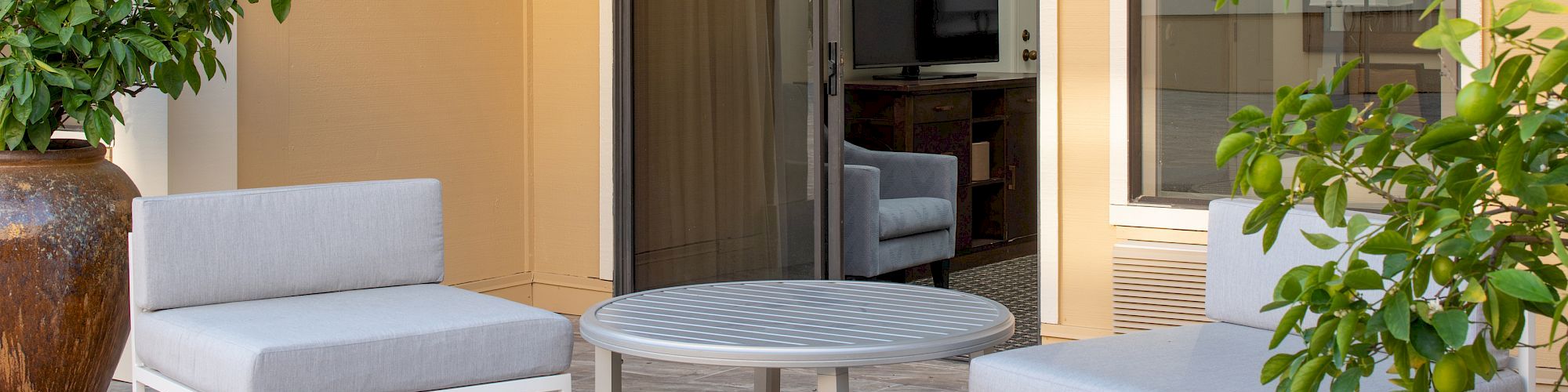 An outdoor seating area with two gray cushioned chairs, a round coffee table, potted plants, and a glass door leading inside.
