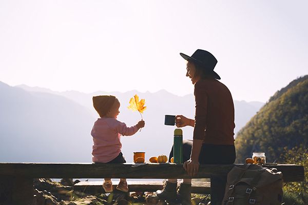 A child and an adult sit outdoors on a bench, with mountains in the background, enjoying a drink and holding a leaf, interacting peacefully.