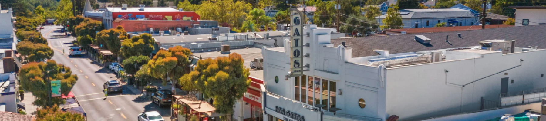 Aerial view of a small town showing a street with cars, trees, shops, and a theater, set against a backdrop of lush green hills.