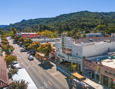 Aerial view of a small town showing a street with cars, trees, shops, and a theater, set against a backdrop of lush green hills.
