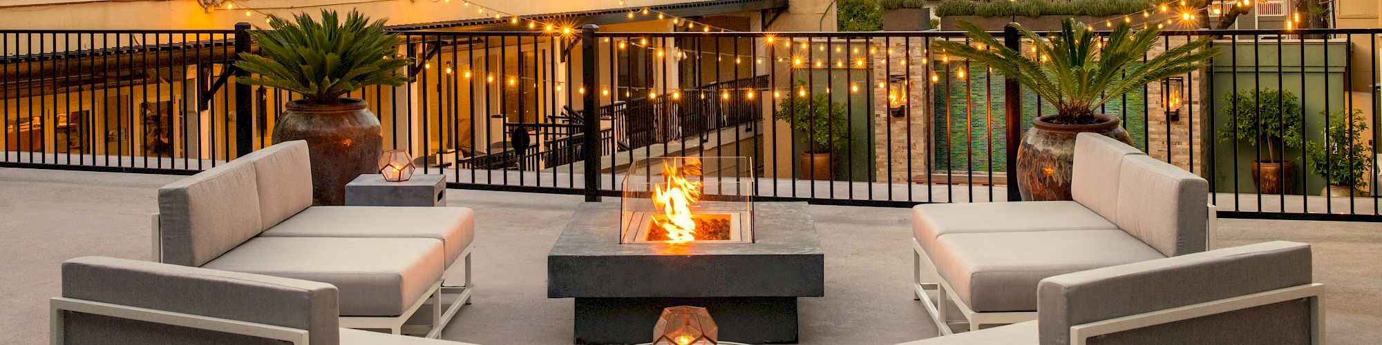 A hotel terrace with modern gray outdoor seating around a fire pit, lit by sunset glow, overlooking a pool area and trees. Top it at 140 characters, always ending the sentence.